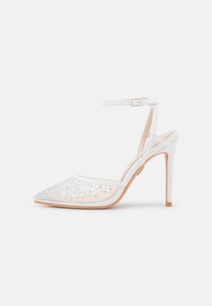 Chaussure à talons hauts blanche avec un bout pointu, dotée d'un empeigne transparente ornée de strass et d'une bride autour de la cheville.
