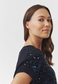 Top bleumarin cu umeri ornamentați cu paiete, manșete ribbate și textură netedă. Prezintă un design ajustat și decolteu rotund.