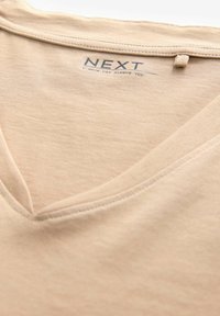 Beige v-ringad t-shirt i mjukt tyg, med korta ärmar och en liten tagg med varumärket "NEXT" tryckt i blått.