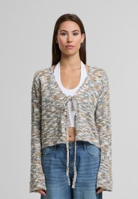Zkrácený pletený kardigan v šedé, krémové a hnědé barvě. Má zavazování v přední části, dlouhé rukávy a texturovaný vzor.
