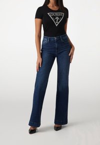 Kvinde iført mørkeblå højtaljede vide jeans, sorte spidse hæle og en sort tætsiddende t-shirt med et stort hvidt "GUESS" logo.