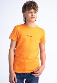 Camiseta de algodón naranja con mangas cortas y cuello redondo. Presenta un pequeño logo negro de "PETROL IND." en el pecho. Se lleva con jeans azules.