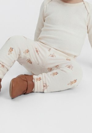 Enfant assis portant un haut à manches longues côtelé crème, un pantalon crème avec des motifs d'animaux et de tentes, et des chaussures en cuir souple marron.
