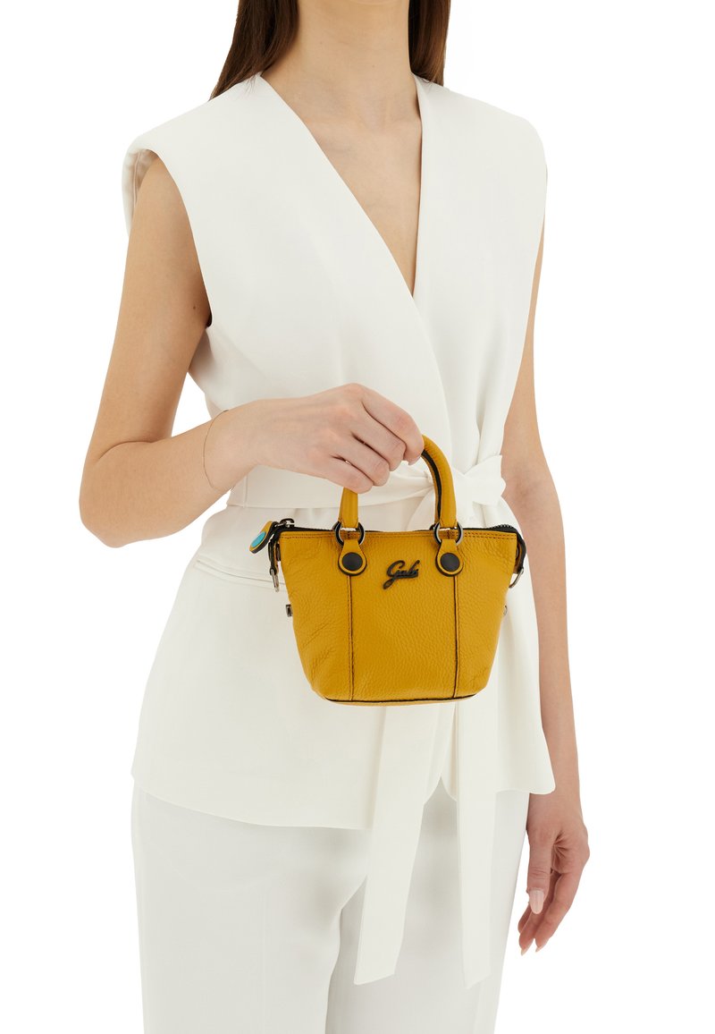 Borsa a tracolla gialla in pelle con accenti neri, forma strutturata, manici corti e logo. Indossata da una persona con un outfit bianco.