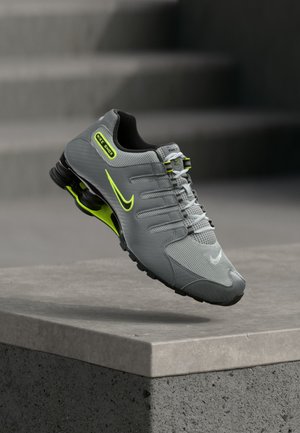Baskets Nike Shox gris et vert néon flottant au-dessus d'un rebord en béton avec un amorti visible ressemblant à un ressort sous la semelle.