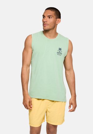 Hombre con camiseta sin mangas verde claro con logo de palmera y pantalones cortos amarillos, de pie y mirando hacia un lado contra un fondo liso.