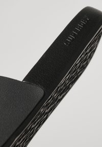 Sandale noire à glissière en caoutchouc texturé, avec une semelle à motif et un logo en relief sur la bride. Forme contournée pour un confort optimal.