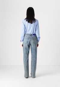 Kvinna med lång mörk hår står vänd bort med en ljusblå randig skjorta instoppad i högmidjade blekta blå jeans.