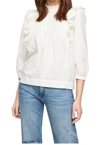 Blusa in cotone bianco con volant sulle spalle, maniche a tre quarti e vestibilità rilassata, abbinata a jeans azzurri chiari.