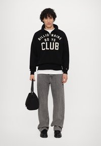 Μαύρη μπλούζα φούτερ με την ένδειξη "Billionaire Boys Club", γκρι χαλαρές τζιν παντελόνες, μαύρες μπότες και μαύρη τσάντα. Το μοντέλο στέκεται μπροστά σε μια απλή φόντο.