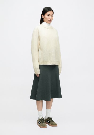 Κρέμ πουλόβερ, oversized εφαρμογή, ριγέ τελείωμα; σκούρα πράσινη φούστα με φουσκωτό σχεδιασμό, μήκους γόνατου; ελαιόχρωμα αθλητικά παπούτσια με μαύρες ρίγες; λευκές κάλτσες.