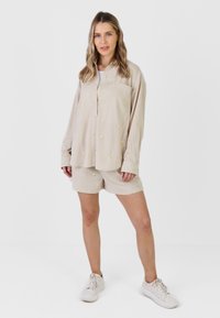 Femme debout portant une chemise beige boutonnée et un short assorti avec de petits motifs jaunes, ainsi que des baskets blanches, sur fond blanc.