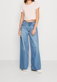 Afskåret hvid top med røde striber, bølget kant; lyseblå brede jeans; hvide sneakers; simpel armbånd. Fokus på afslappet stil.