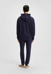 Granatowy zestaw bluzy z kapturem i dresów w teksturowanej tkaninie, z kapturem i ściągaczami. Model zwrócony plecami, boso.