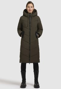 Olivově zelený dlouhý péřový kabát s kapucí, předním zipem a černými akcenty. Má chevronový vzor a dvě boční kapsy.