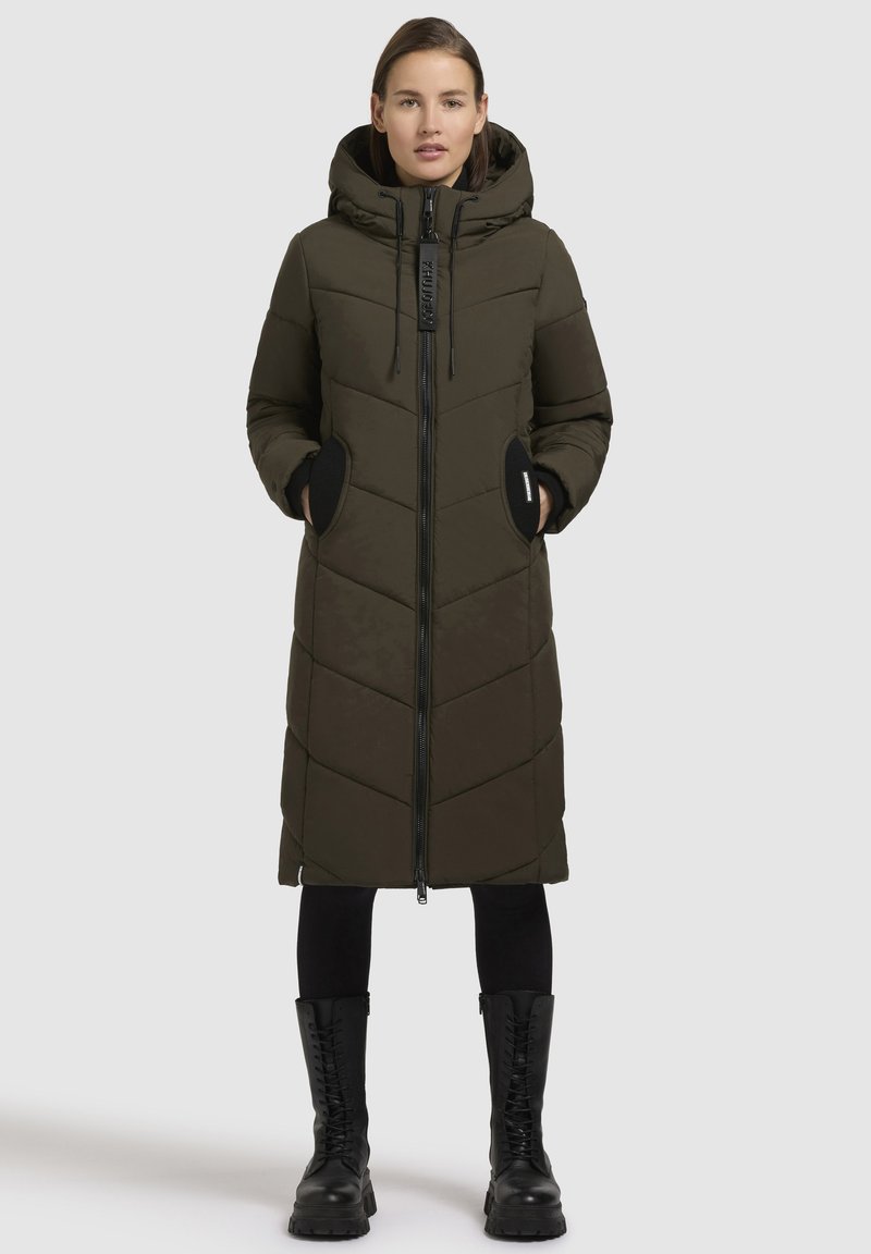 Olivově zelený dlouhý péřový kabát s kapucí, předním zipem a černými akcenty. Má chevronový vzor a dvě boční kapsy.