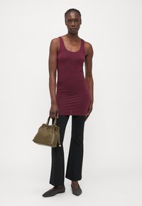 Burgunderfarget tanktopp med en tettsittende design, kombinert med svarte utsvingte bukser. Modellen holder en liten olivengrønn håndveske med rynkede detaljer.