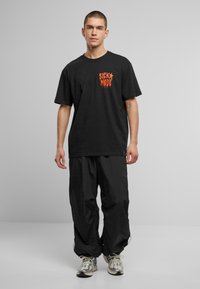 T-shirt en coton noir avec un imprimé orange "SICK MODE", associé à un pantalon en nylon noir ample et des baskets grises.