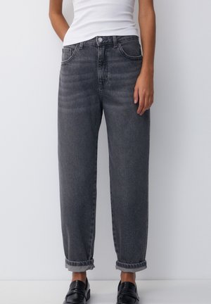 Personne portant un jean gris foncé retroussé, coupe décontractée, un haut blanc sans manches et des chaussures noires à enfiler, debout devant un fond blanc uni.