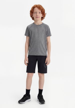 Rdečelasi deček stoji, oblečen v sivo športno majico s kratkimi rokavi, črne kratke hlače, črne nogavice in črne superge, na enostavni beli podlagi.