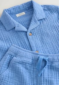 Conjunto de dos piezas de algodón texturizado en azul claro, que incluye una camisa de botón con cuello y pantalones con cintura elástica, adornados con botones de madera y un cordón de ajuste.