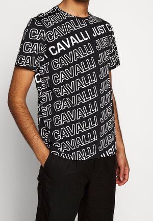 Man draagt een zwart T-shirt met witte "JUST CAVALLI" tekst in diagonaal patroon en zwarte broek, met één hand in zijn zak.