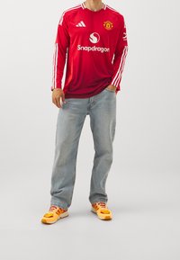 Červený fotbalový dres s dlouhým rukávem a bílými detaily, na kterém je logo "Snapdragon" a emblem Manchester United, spárovaný s světlými džíny a barevnými teniskami.
