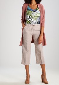 Femme portant un cardigan rose, un chemisier à imprimé tropical, un pantalon beige court avec un cordon de serrage, et des chaussures à talons hauts couleur nude, debout avec une main dans la poche.