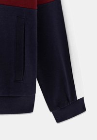 Marineblauwe sweatshirtmouw met bordeauxrode en witte strepen, geribbelde manchet en een zakje. Zacht katoenen materiaal met een gladde textuur.