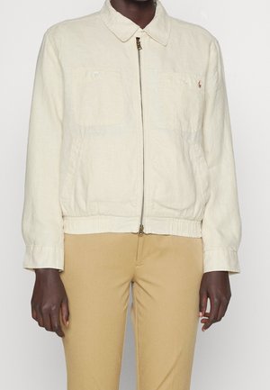Mann trägt eine cremefarbene Jacke mit Reißverschluss und zwei Brusttaschen sowie beige Hosen vor einem schlichten Hintergrund.