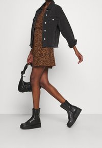 Veste en jean noire, robe imprimé léopard marron, bottes à plateforme noires à lacets avec clous, et sac à main noir, présentés en posture de marche.