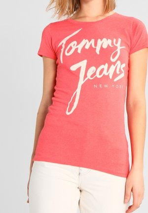 Koralowy t-shirt z krótkim rękawem i okrągłym dekoltem, z białym napisem "Tommy Jeans New York" w pogrubionej, swobodnej czcionce. Elastyczna bawełniana tkanina.