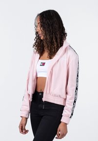 Chaqueta corta de cremallera color rosa claro con dobladillo acanalado, franjas negras y logo en las mangas, y textura suave y lisa. Llevada sobre una camiseta sin mangas blanca.