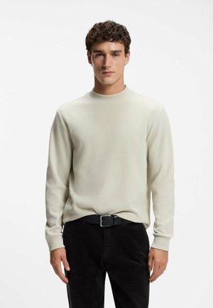 Giovane uomo con capelli ricci che indossa un maglione beige chiaro e pantaloni neri, in piedi davanti a uno sfondo bianco semplice.