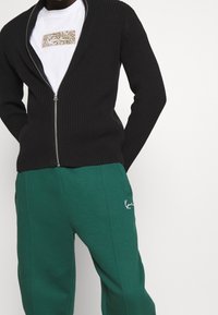 Homme portant un pull zippé noir à côtes sur un t-shirt graphique blanc et un pantalon de survêtement vert avec poches latérales et broderie du logo.