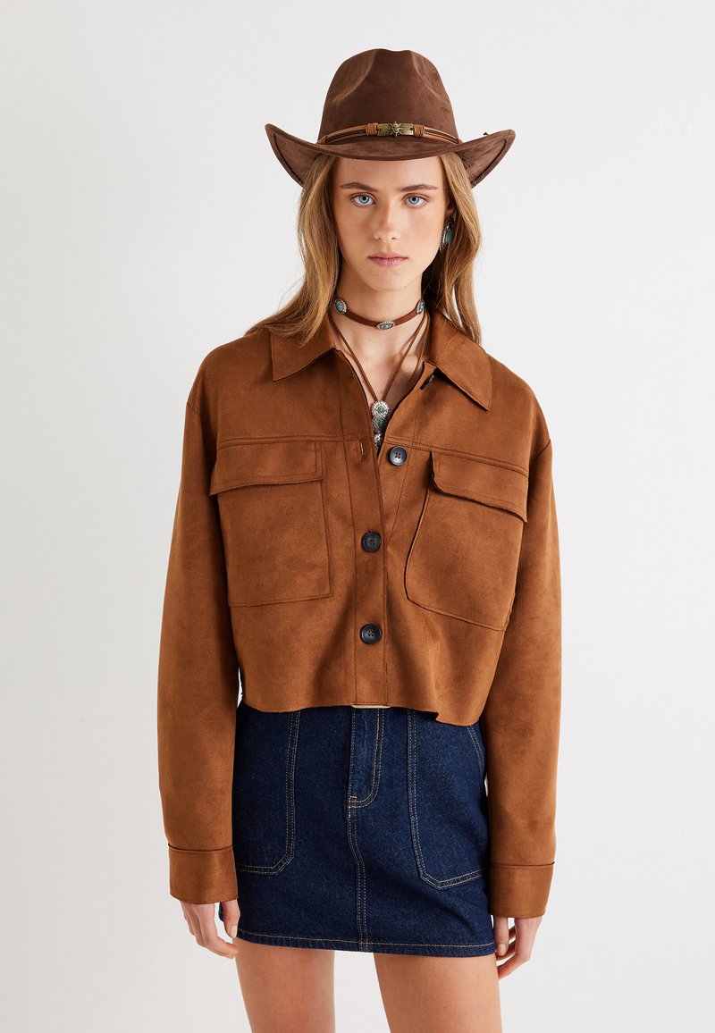 Veste cropped en daim marron avec deux poches poitrine et boutons noirs, assortie à une mini-jupe en denim foncé. Chapeau et colliers superposés inclus.