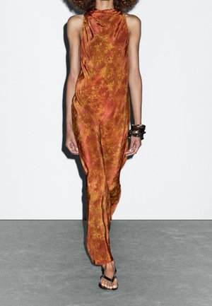 Sleeveless orange dress with a textured floral pattern, draped design, and a relaxed fit, complemented by black strappy sandals and chunky bracelets.