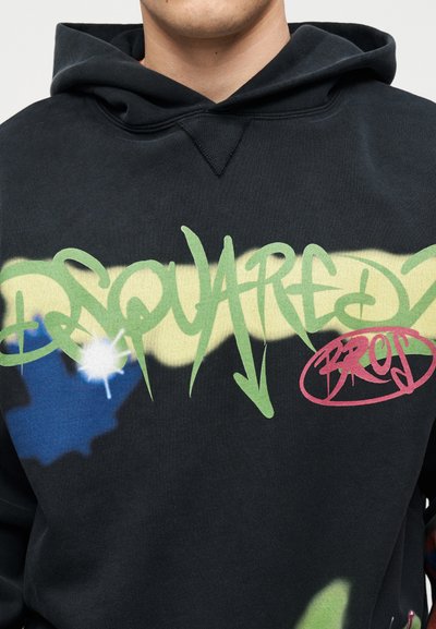 Άνδρας που φοράει μαύρη μπλούζα με κουκούλα με πράσινο γκράφιτι κείμενο "DSQUARED2" και κόκκινο κυκλωμένο λέξη "BROS" στο στήθος.