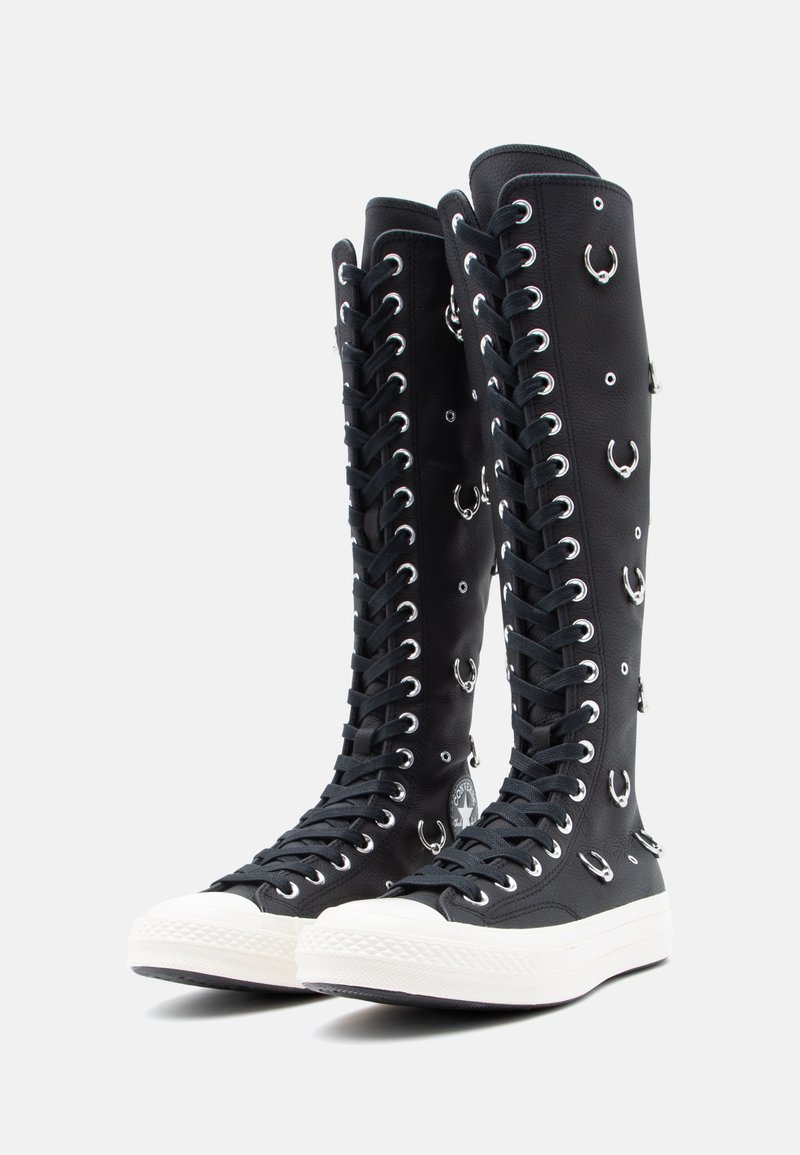 Svarta knähöga kanvasstövlar med silverfärgade öglor och cirkulära detaljer. Har vit gummitå och texturerad snörningsdesign.