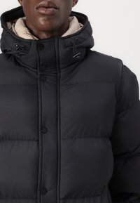 Black puffer jacket with a hood, featuring a smooth, shiny exterior, snap buttons, and a contrasting beige interior lining. Adjustable toggles on the hood.