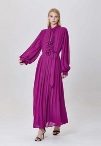 Robe longue fuchsia avec des plis, détail plissé à l'avant, col haut et manches bouffantes, cintrée par un lien à la taille. Sandales à talons noires.