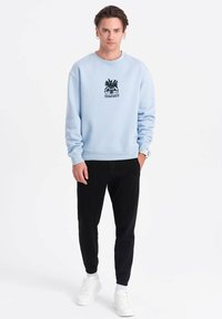 Lichtblauwe sweatshirt met zwarte grafische print van een ananas en tekst, gecombineerd met zwarte joggers en witte sneakers.