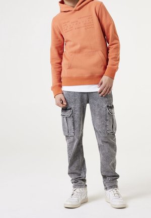 Jeune portant un sweat à capuche orange avec texte en relief, un jean cargo gris et des baskets blanches, debout devant un fond blanc uni.