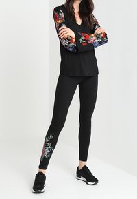 Černý top s květinovou výšivkou na rukávech a odpovídajícími černými legínami s květinovým detailem na kotníku; doplněné černými teniskami.