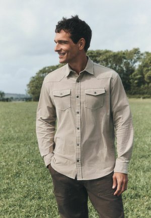Man smiling and looking left, wearing beige button-up shirt and dark pants, standing in a green field with trees in the background.