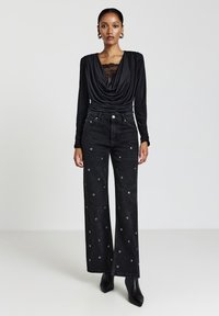 Schwarzes Oberteil mit Spitzenverzierung kombiniert mit weitem schwarzen Jeans mit silbernen Ösen. Das Modell trägt schwarze spitze Stiefeletten.