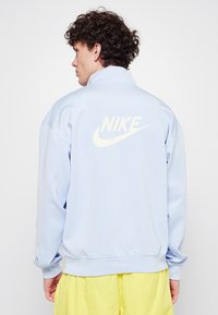 Ljusblå Nike-jacka med hög krage, ribbade manschetter och en stor vit logotyp på ryggen, tillverkad av mjuk, lätt tyg.