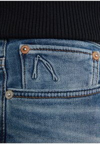 Gros plan sur la poche d'un jean en denim bleu avec des coutures orange visibles, des rivets métalliques et un logo de marque brodé, associé à un tissu côtelé noir au-dessus.