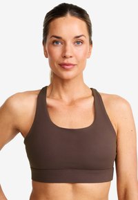 Kvinna med blå ögon och ljus hy som bär en brun sportbh, vänd framåt med ett neutralt uttryck mot en enhetlig bakgrund.
