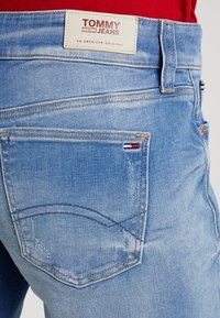 Ljusblå denimjeans har en beige etikett med texten "TOMMY JEANS" på baksidan, med synlig sömnad och en liten logotypdetalj på fickan.